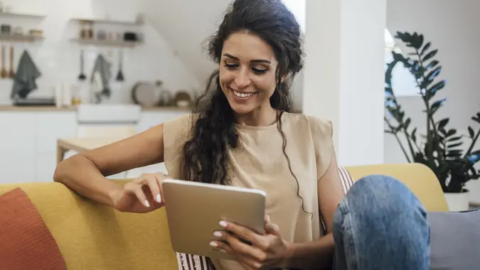 Happy young woman sitting on a couch outside of her kitchen and using a tablet next to a plant in her home