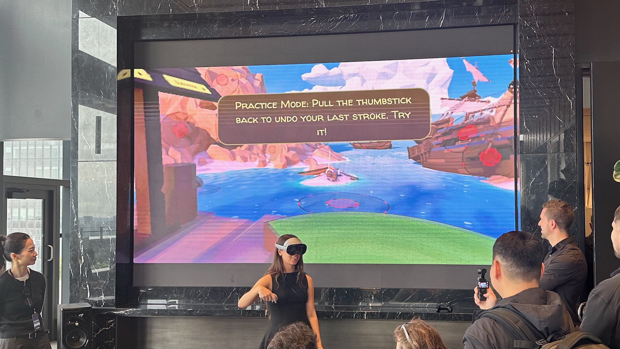 A photo of a Samsung engineer playing Walkabout Mini Golf while wearing the Galaxy XR headset and using a Galaxy XR controller in her right hand, a screen showing the game behind her.