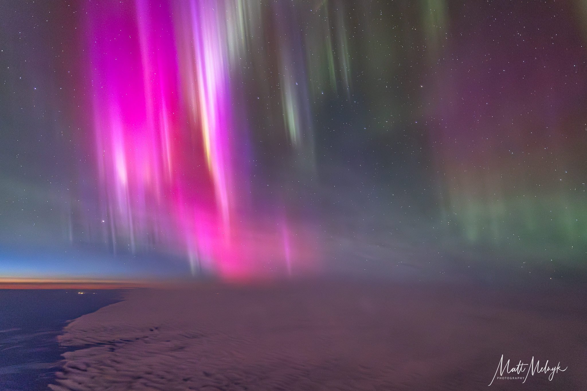 curtains of magenta and green light dance in the sky, a thin blue line marking Earth's atmosphere is visible in the distance. 