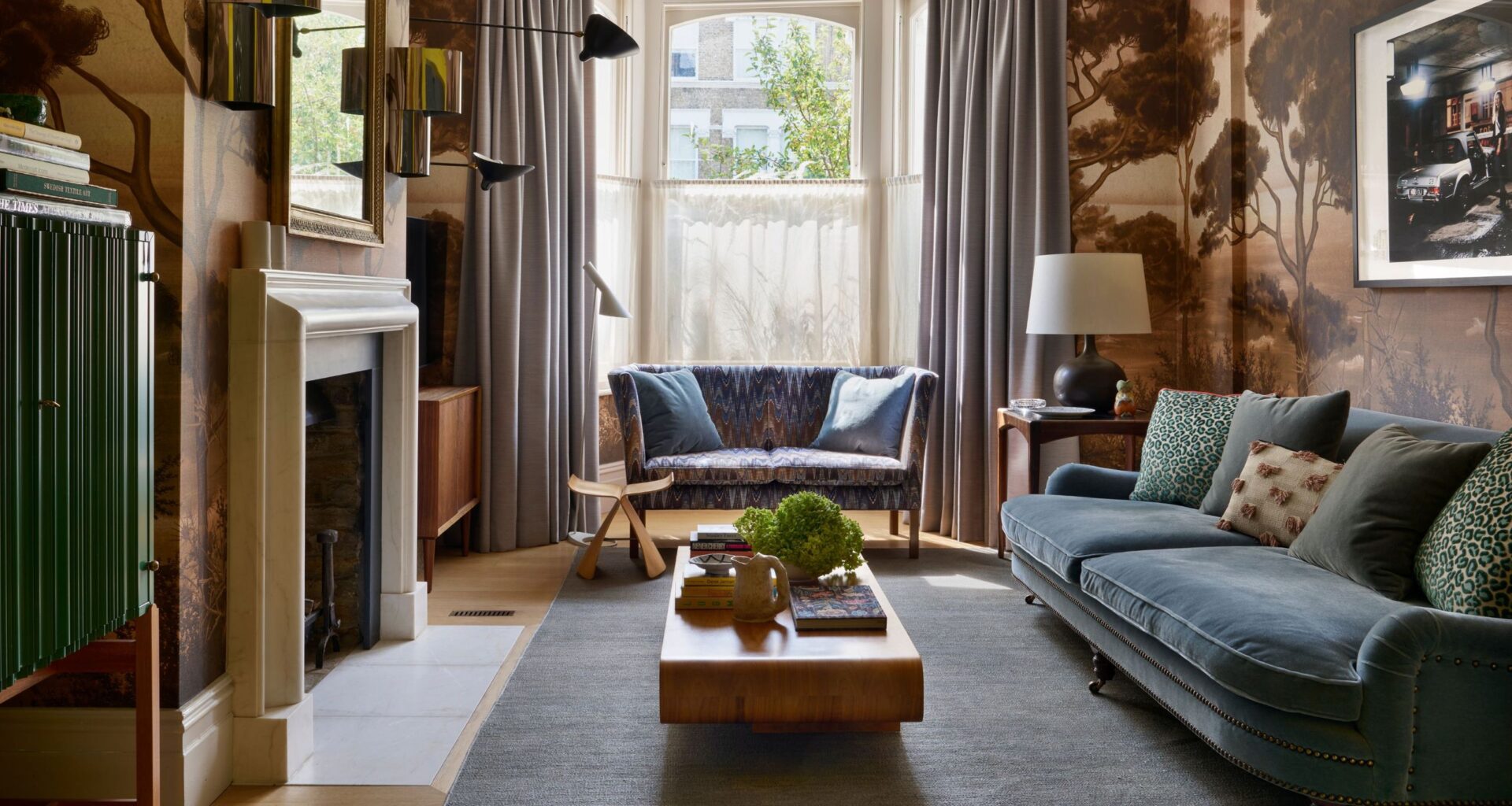 A living room area with a large sofa and a smaller sofa beside the window. On either side of the sofa are curtains hanging. In the middle of the room is a coffee table with a plant on top of it as well as other coffee table essentials. The living room also features a fireplace as well as a mirror and artwork.