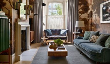 A living room area with a large sofa and a smaller sofa beside the window. On either side of the sofa are curtains hanging. In the middle of the room is a coffee table with a plant on top of it as well as other coffee table essentials. The living room also features a fireplace as well as a mirror and artwork.