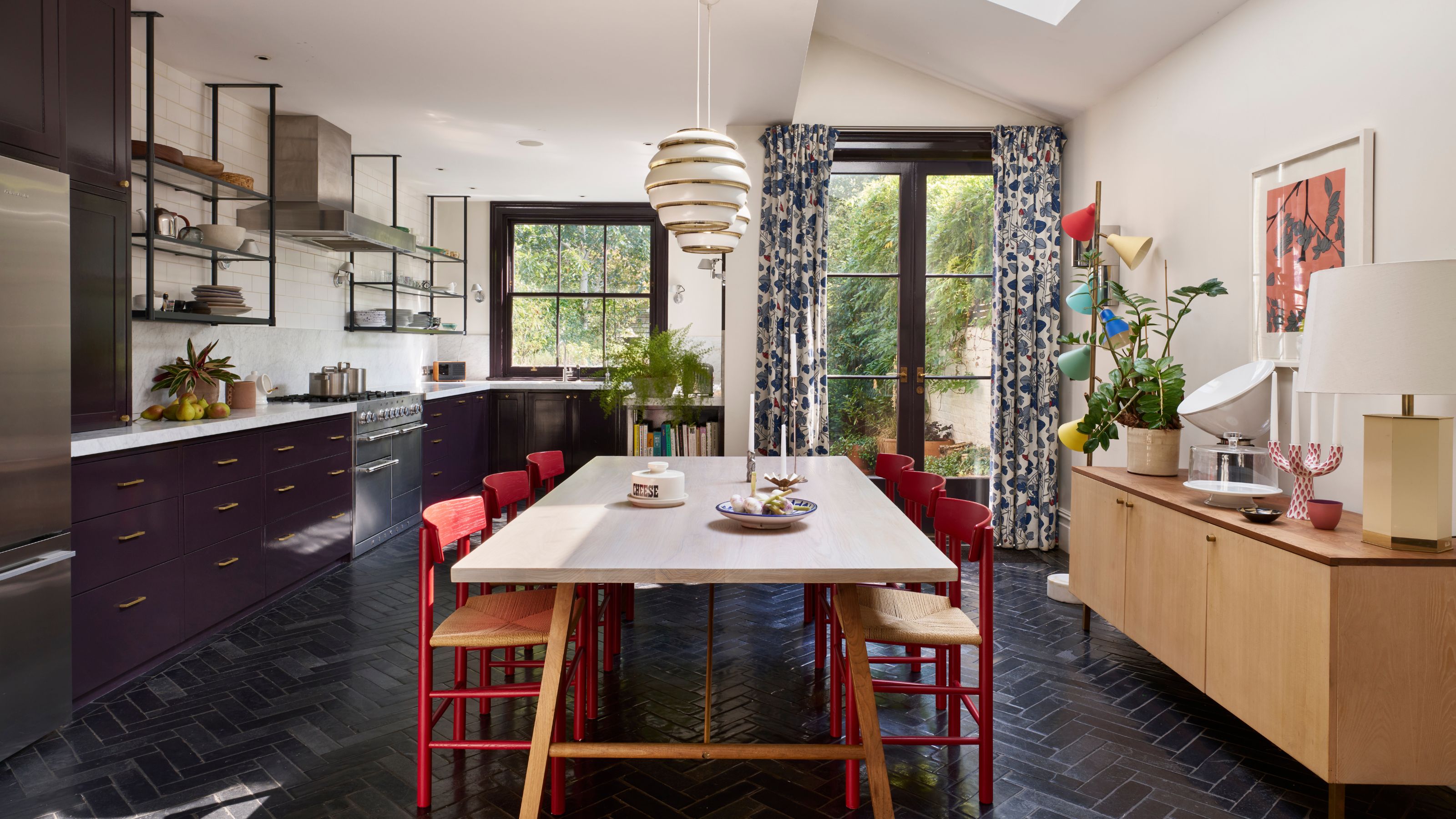 A kitchen with an open plan layout that also has a dining area. In the kitchen are dark cabinets with open shelving above the cabinets as well as herringbone floor tiles.