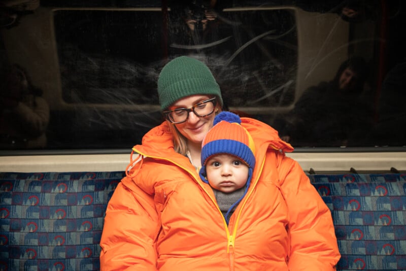 A woman in glasses and a green beanie sits on a train, wearing an orange puffer jacket. A baby in a blue and orange hat peeks out from inside her jacket, looking at the camera.