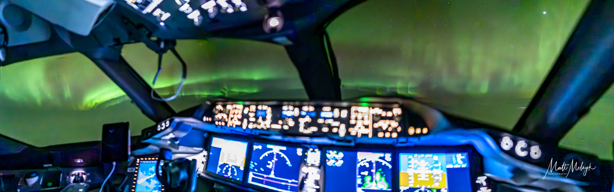 inside airplane cockpit shows lights with controls and large windows with nothing but green auroras stretching outside.