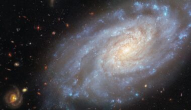 spiral galaxy occupies most of the image. It is a slightly tilted disc of stars, yellow-white in the centre and blue in the outskirts, showing light from different stars in the galaxy. Its spiral arms curl outwards from the centre, speckled with blue star clusters. Dark reddish threads of dust swirl around the galaxy&rsquo;s centre. The backdrop is two medium-sized and many small, distant galaxies on a black background.