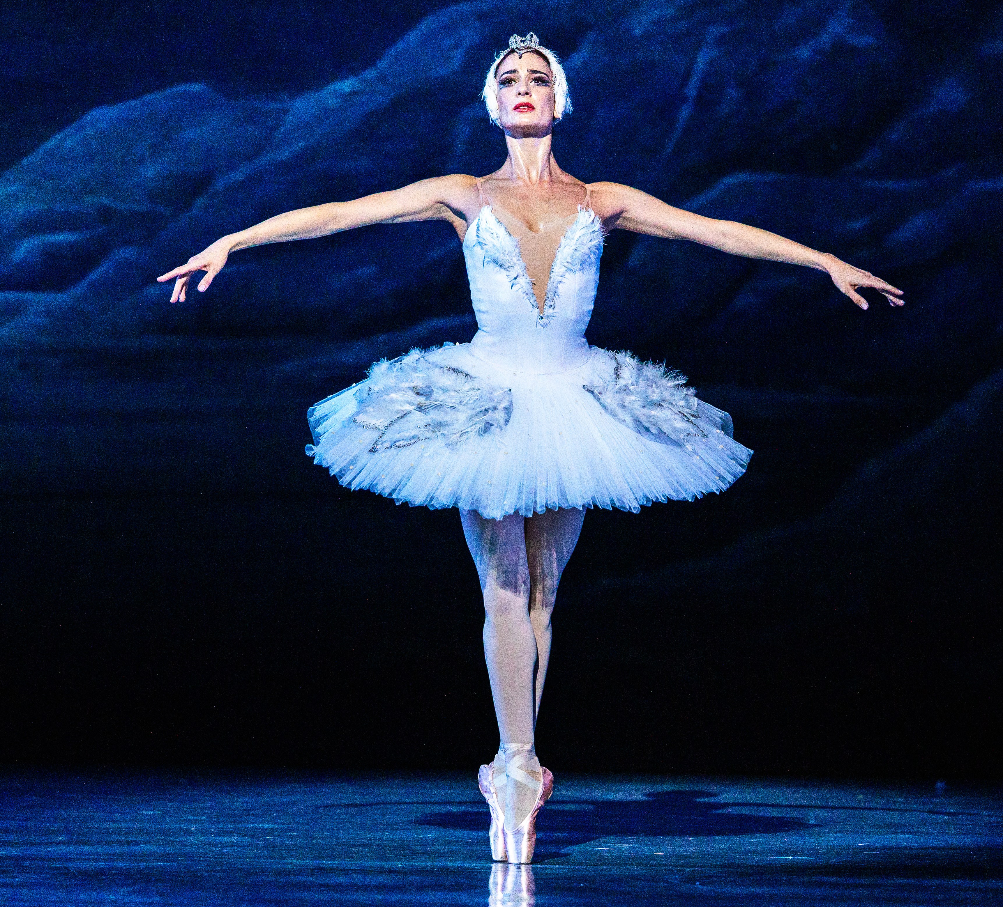 A ballet dancer dressed as a swan with a white feathered tutu and tiara poses on stage with arms extended.