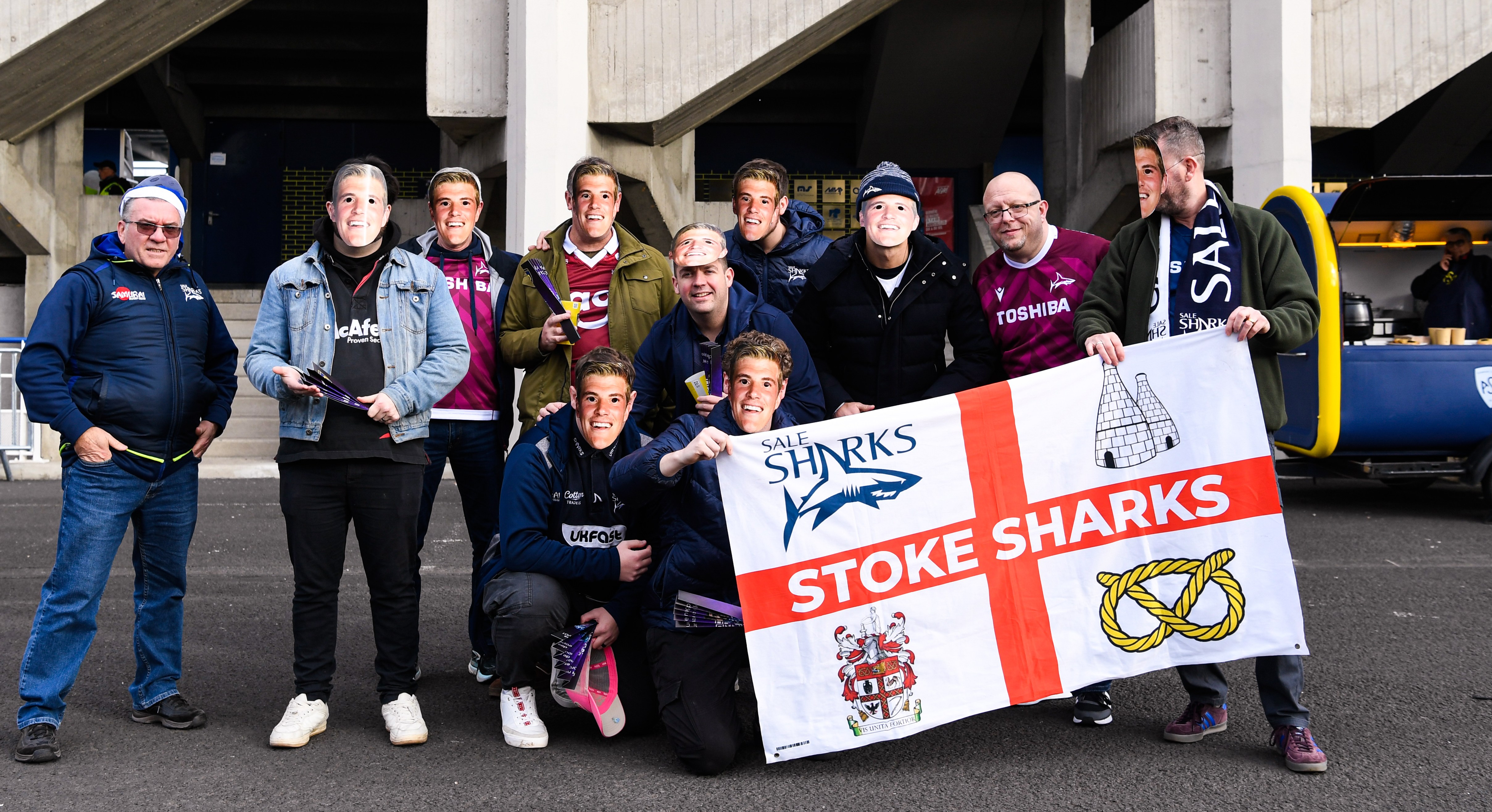 Fans of Sale Sharks pose with a mask of Ernst VAN RHYN, captain of Sale Sharks, holding a "Stoke Sharks" flag.