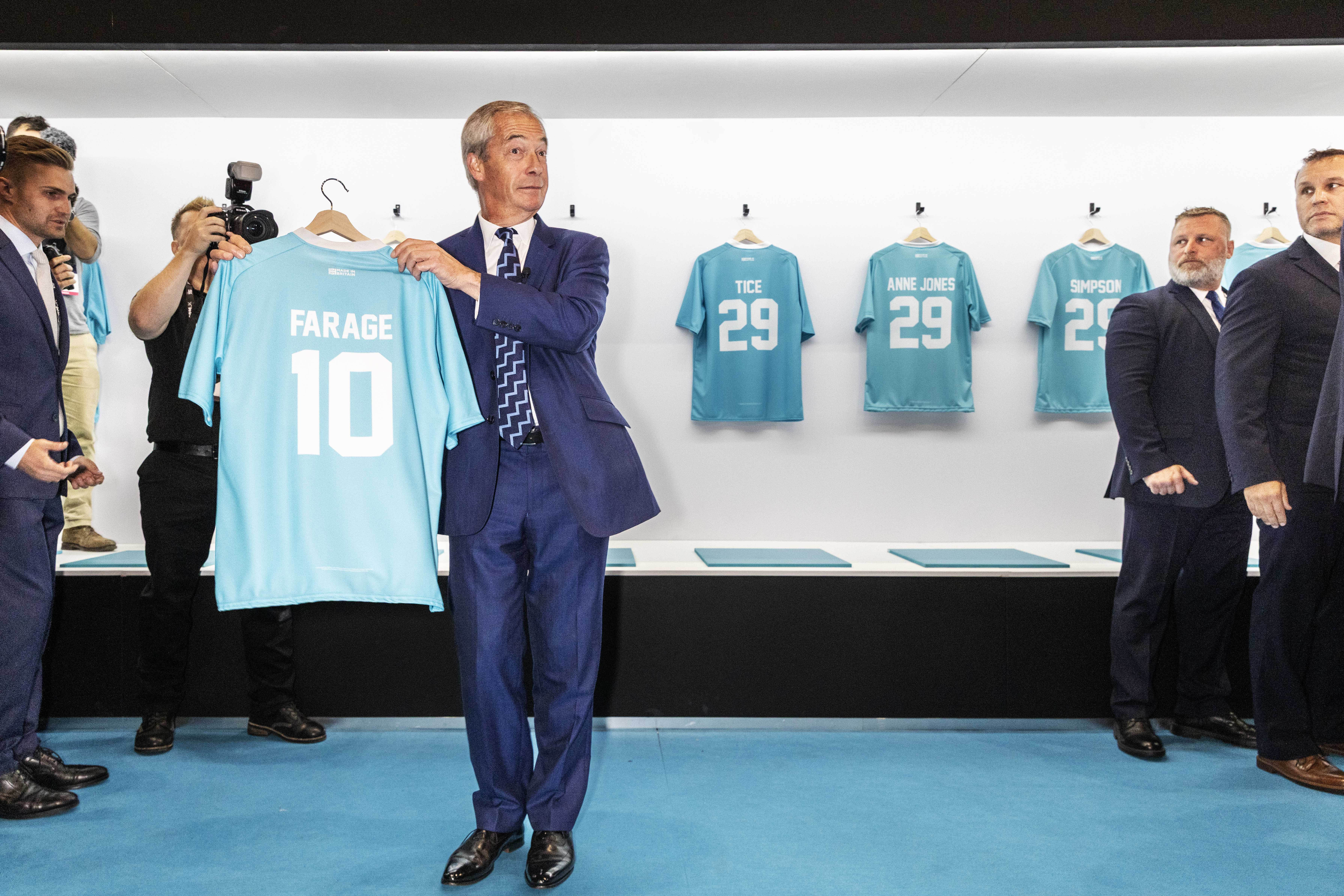 Nigel Farage holding up a light blue shirt with the name "FARAGE" and the number "10" on the back, while other shirts are hanging on the wall behind him.