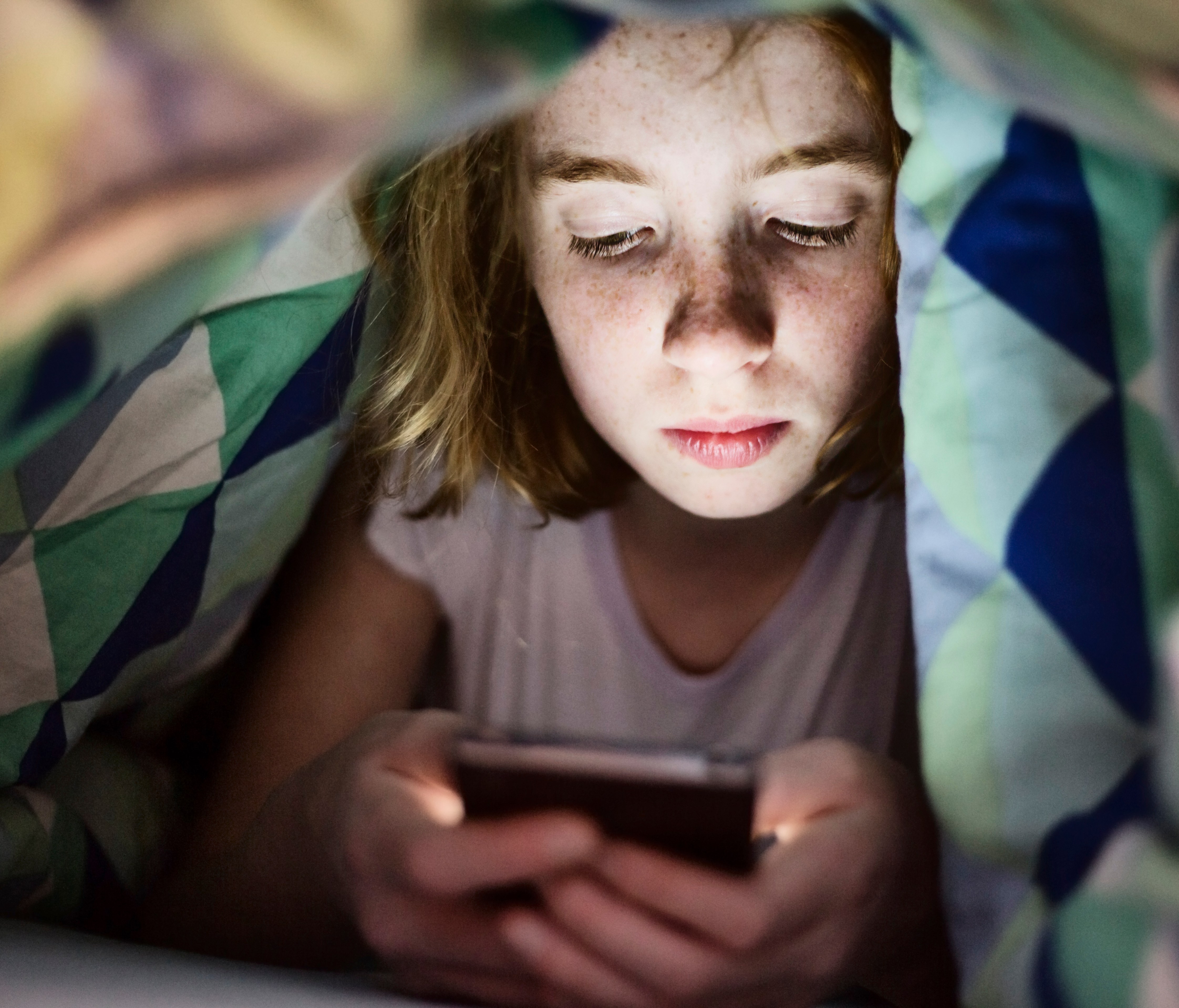 Girl lying under blanket in bed looking at her smartphone