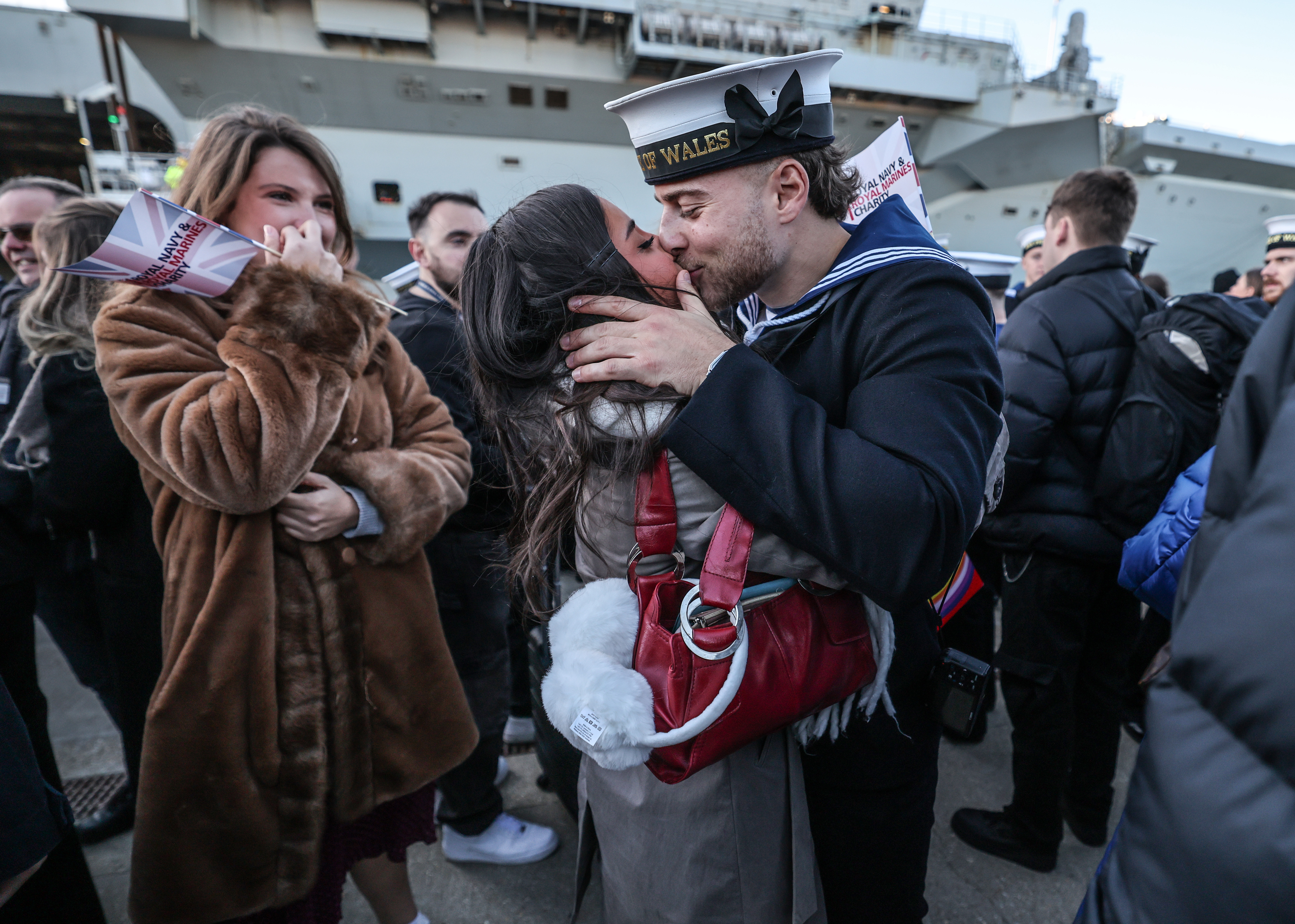 A Royal Navy sailor in uniform kisses his loved one on a quayside after returning from deployment.