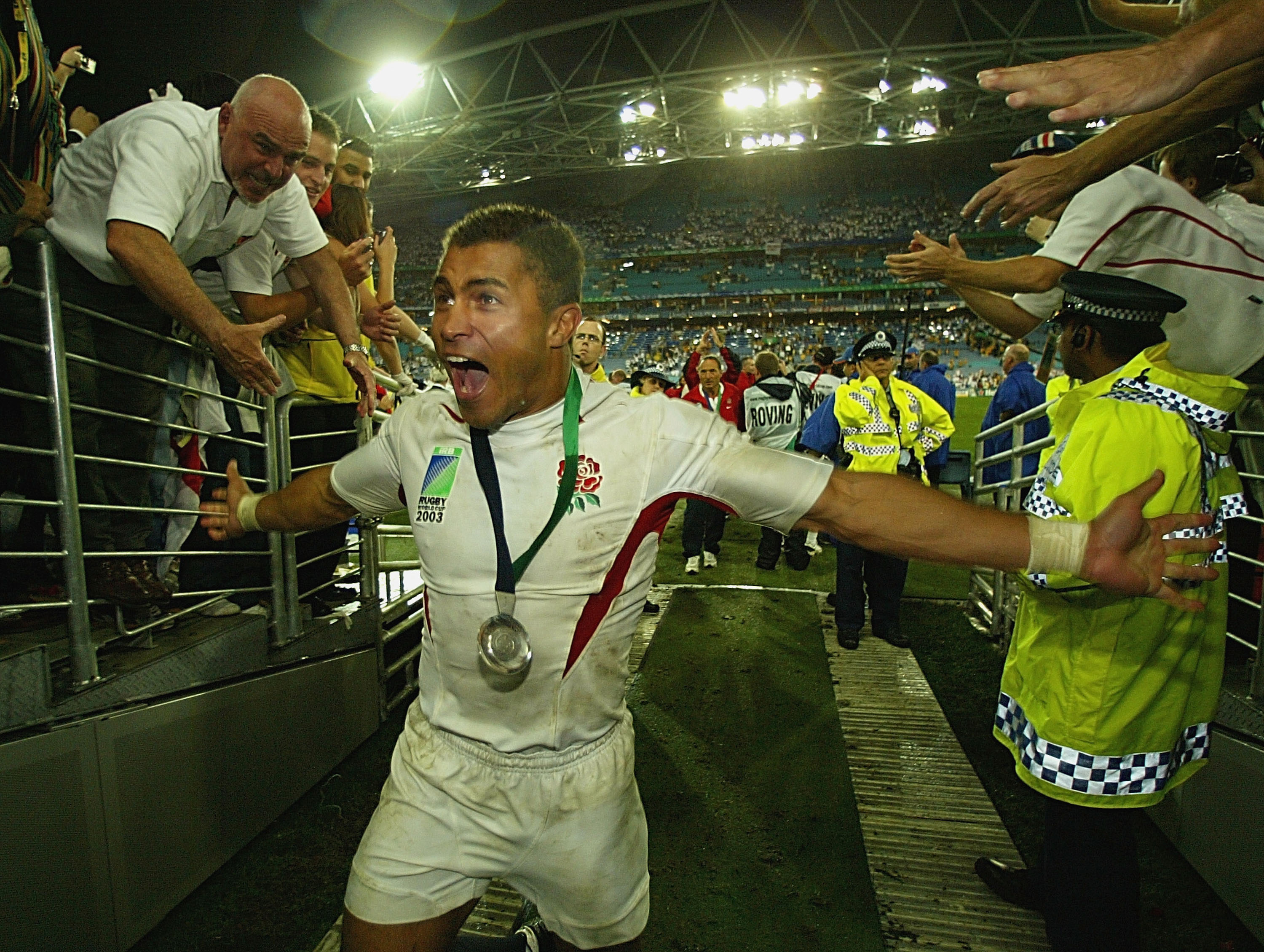 Rugby player Jason Robinson, in a white and red uniform with a medal around his neck, cheering and reaching out to the crowd.