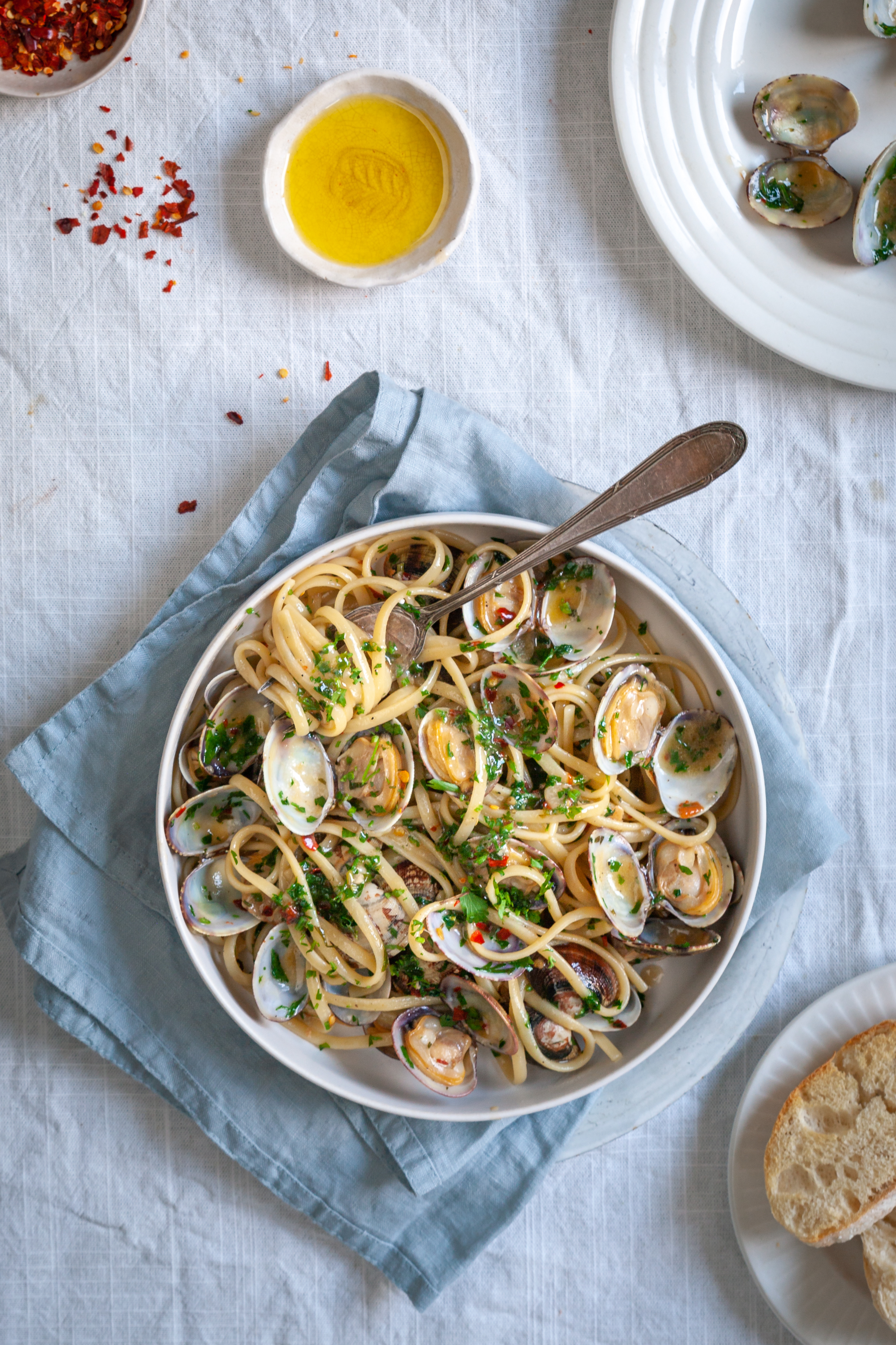 A bowl of pasta vongole on a white tablecloth, garnished with red chili flakes and a side of olive oil.