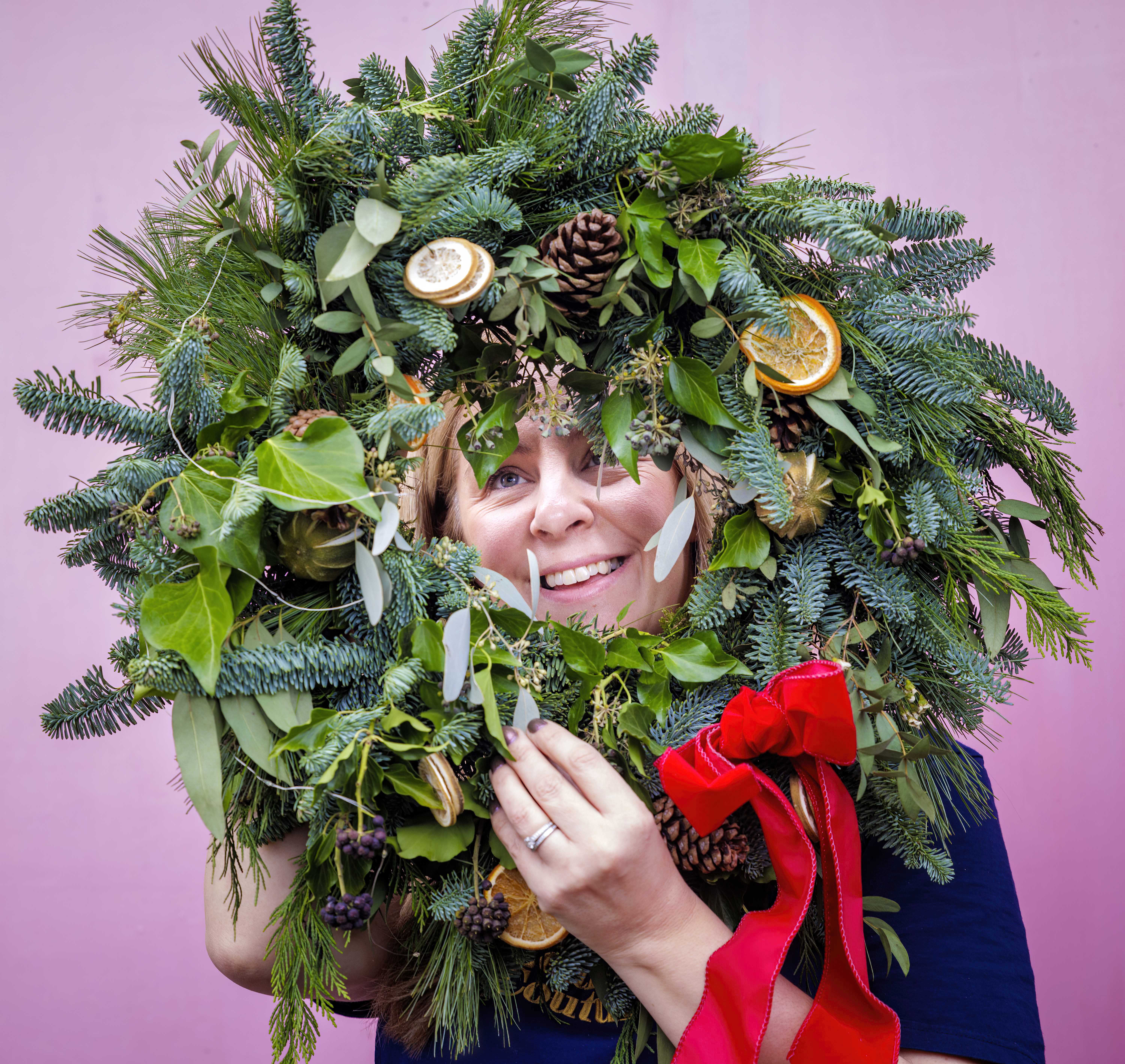 Sarah Richardson of Leafy Couture poses with a Christmas wreath against a pink background.