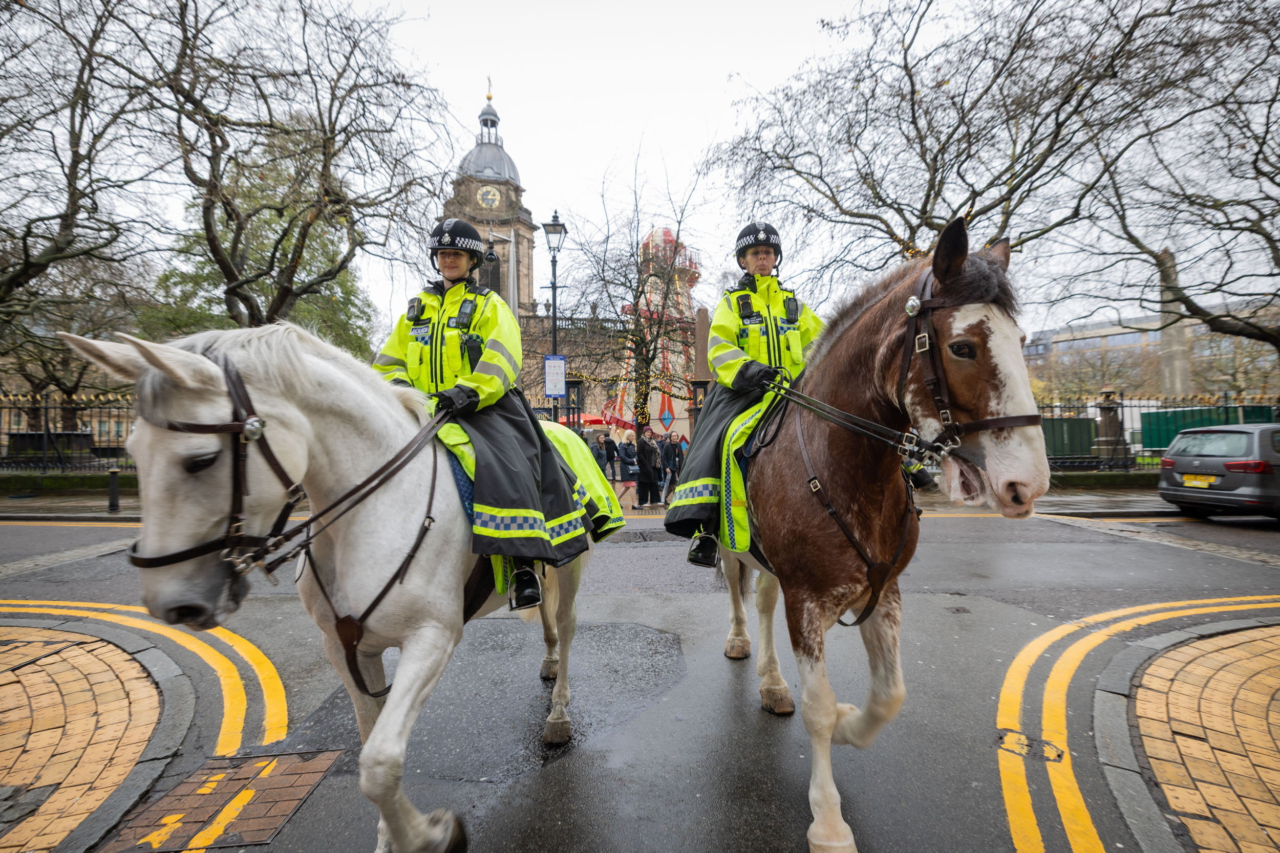 Two police officers in bright yellow high-visibility jackets and helmets ride a white and a brown horse on a wet street.