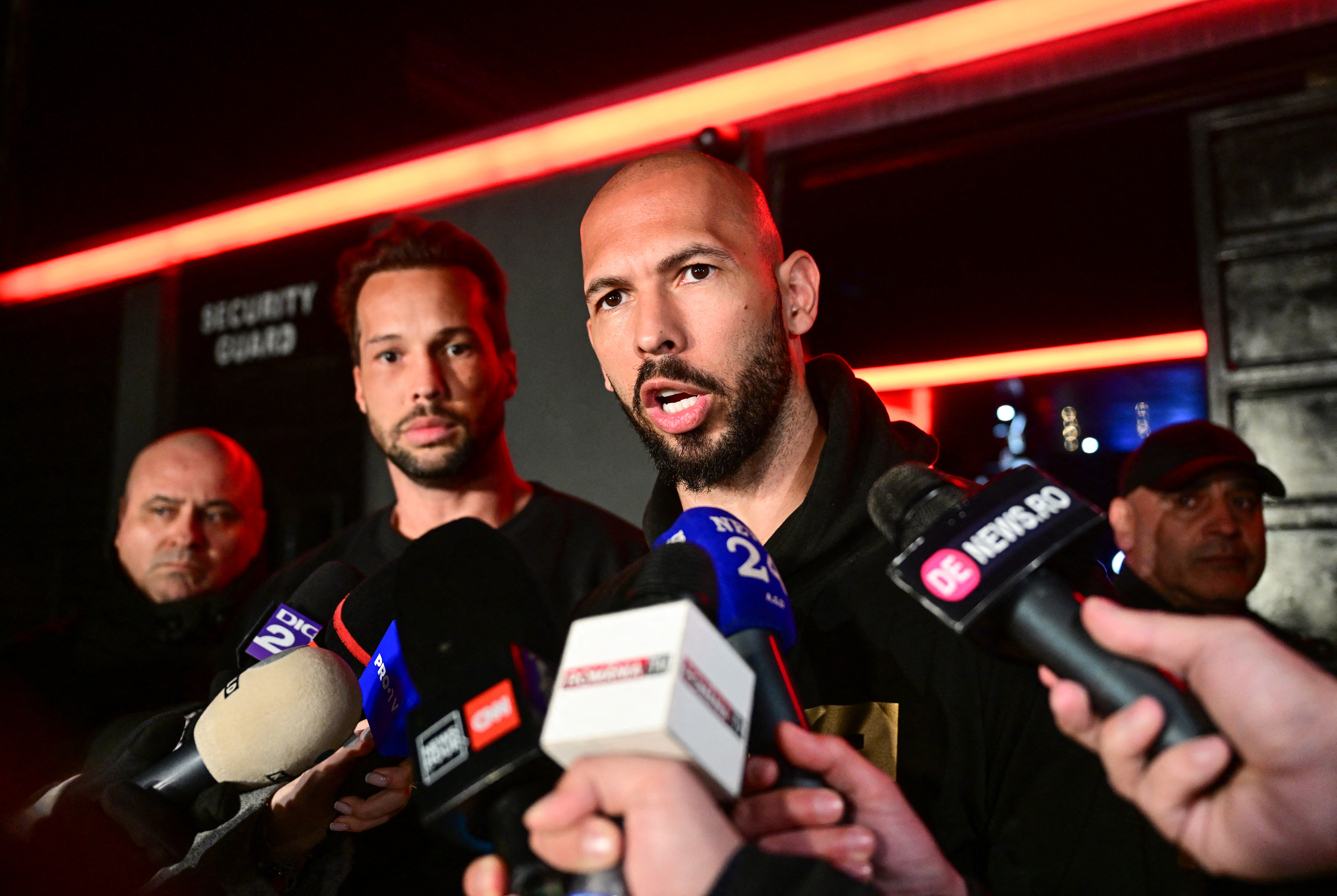 Andrew Tate speaks into multiple microphones with his brother Tristan Tate standing behind him.