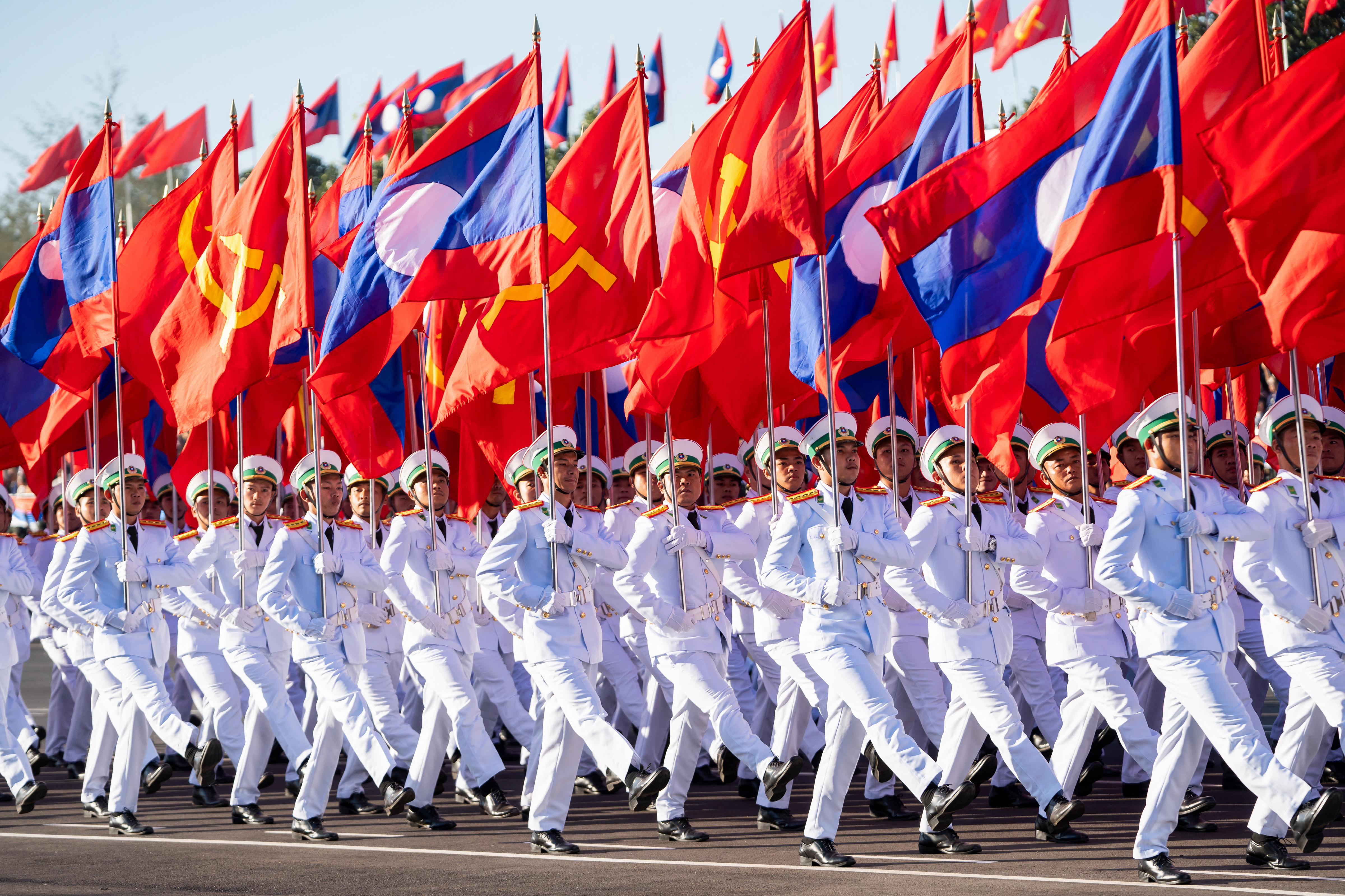 Laotian service members in white uniforms march with Lao National Day flags and communist flags.