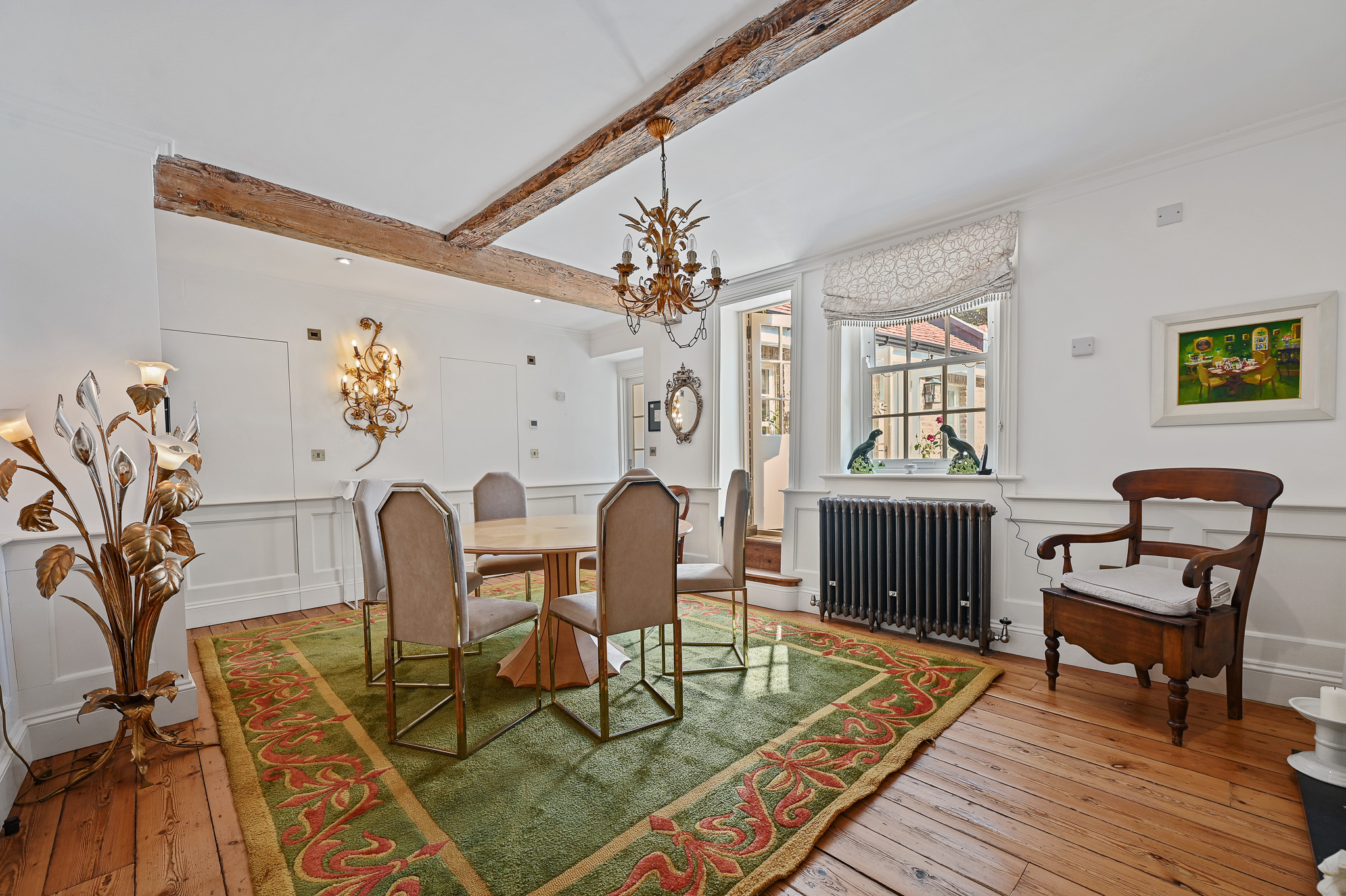 A dining room with white walls, exposed wooden beams on the ceiling, a round dining table, and a green rug with a red pattern.