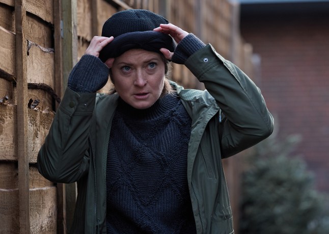 Becky Swain looking suspicious as she lurks by a fence in Coronation Street, wearing a navy hat and jumper.