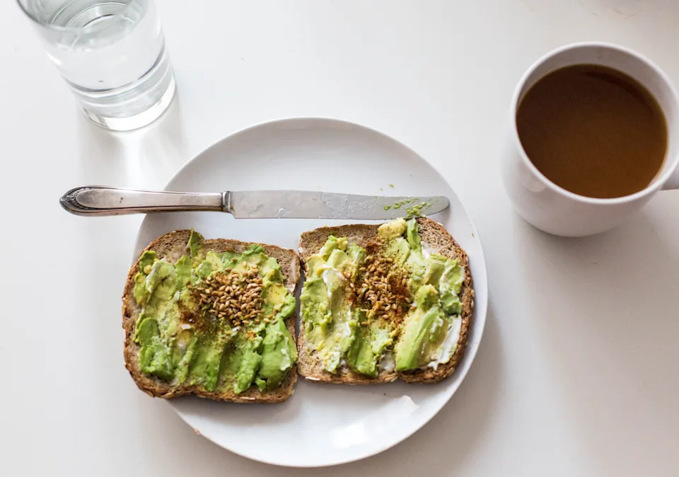 Plate with avocado toast, topped with seeds, beside a glass of water and a cup of coffee on a table