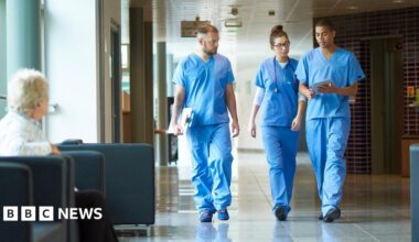 Three medics in blue scrubs, two men and a woman, walk along a hospital corridor. One of the men is holding a clipboard, the other some papers and the woman has a stethoscope around her neck. They are walking side by side, engaged in conversation. In the foreground, there is woman sitting on a black cushioned chair. She is turned away from the camera, looking towards the medics.