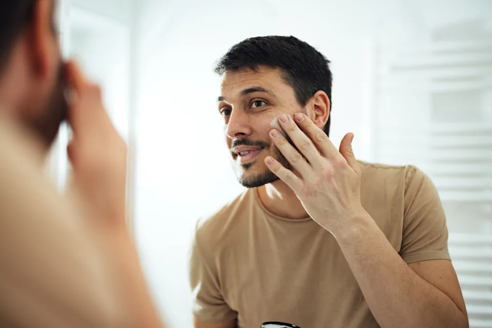 Handsome man standing in the bathroom and looking at the mirror while applying SPF