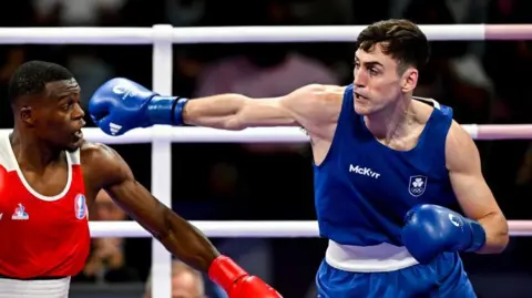 Sportsfile/Getty Two men are boxing in a ring. The man on the left is black and dressed in red and white kit and gloves, the man on the right is white and dressed in blue and white kit and gloves. The background is out of focus, but there is an audience behind them and a male referee in a white shirt and black bow tie. 