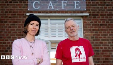 Emma Fernandez and Patrick Matthews standing in front of the lido cafe. Emma wears a pink cardigan and a black beanie hat, and Patrick wears a red t-shirt.