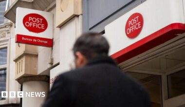 A man in a dark coat walks by a Post Office branch