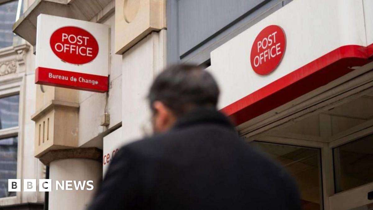 A man in a dark coat walks by a Post Office branch