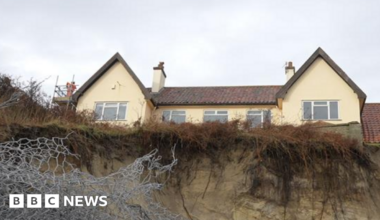 A view from the bottom of a cliff looking up toward a house that can be seen towering over. It is a yellow house with several windows and two chimneys. Only the top storey of the house can be seen. Metal wires can be seen toward the bottom left of the image as part of sea defences.