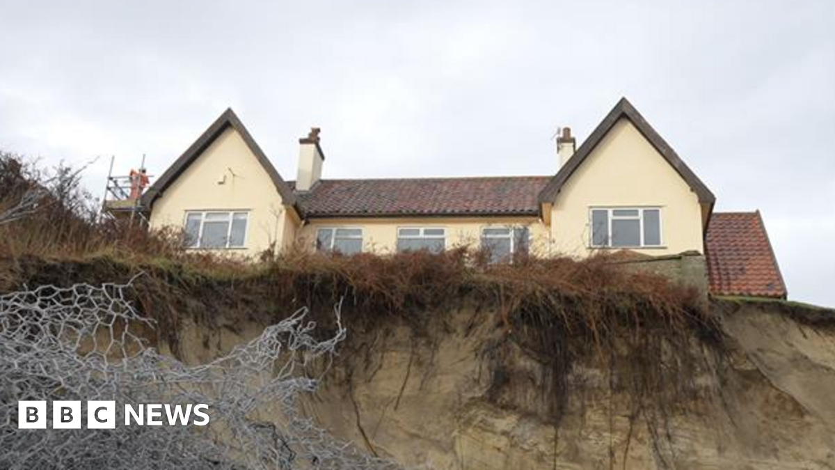 A view from the bottom of a cliff looking up toward a house that can be seen towering over. It is a yellow house with several windows and two chimneys. Only the top storey of the house can be seen. Metal wires can be seen toward the bottom left of the image as part of sea defences.