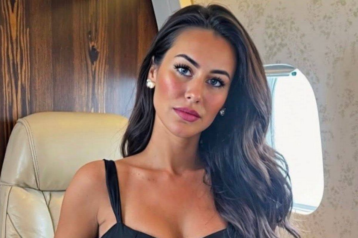 An individual with long hair is seated on a chair inside an aircraft, with a television screen visible in the background.