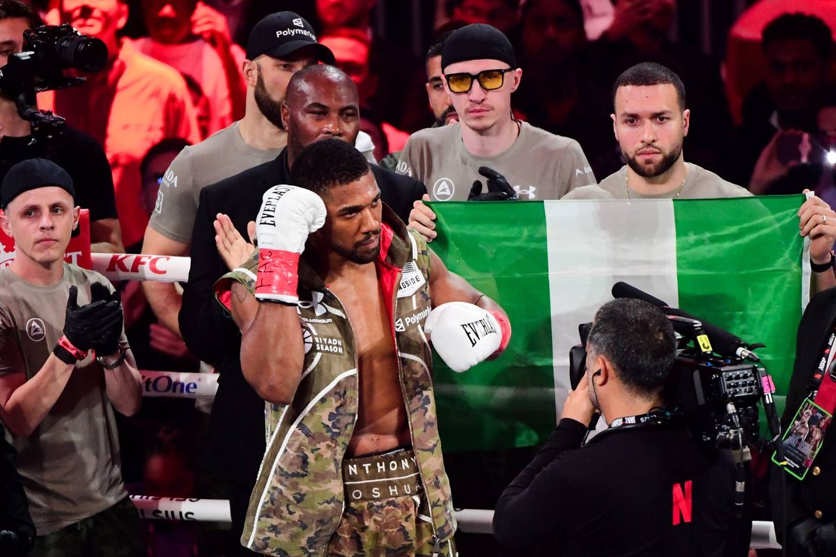 A male boxer, adorned in traditional fight attire, stands prominently in the center of the ring. He holds his hands up in a gesture of victory or acknowledgment, with a national flag draped over his shoulders. Behind him, several individuals, including what appears to be his coach or team members, are gathered, attentively observing the scene. The atmosphere is charged with anticipation, capturing a moment of triumph or recognition within a competitive setting.