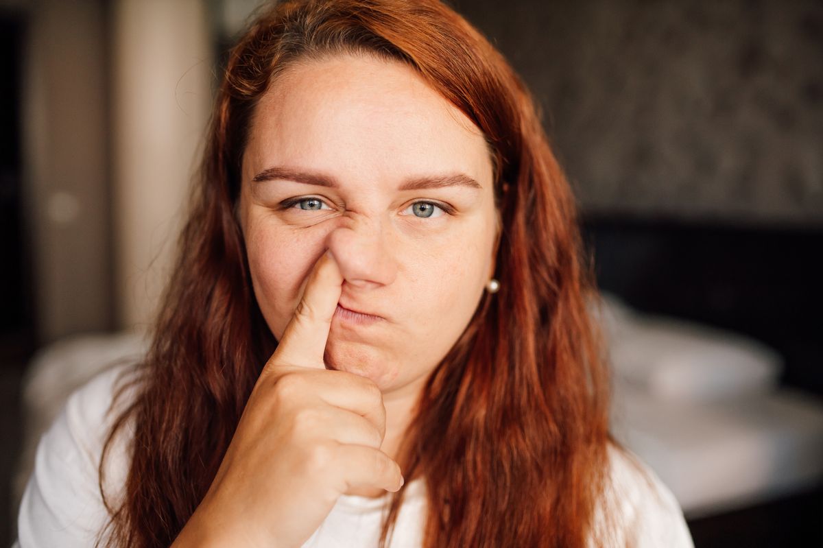 Close-up image of mature redhead woman in white T-shirt standing in cozy bedroom engrossed in act of picking her nose. Woman facial expression invites viewers to ponder importance of knowing good manners and etiquette