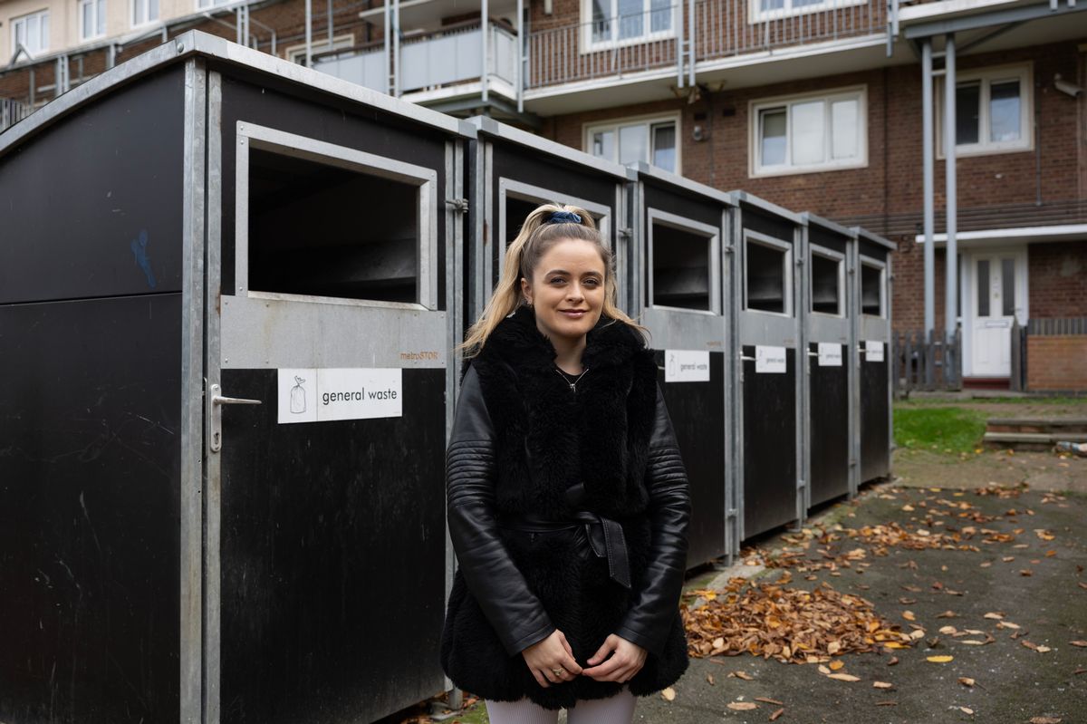 Loretta Alvarez standing in front of a row of black rubbish and recycling bins outside a block of flats