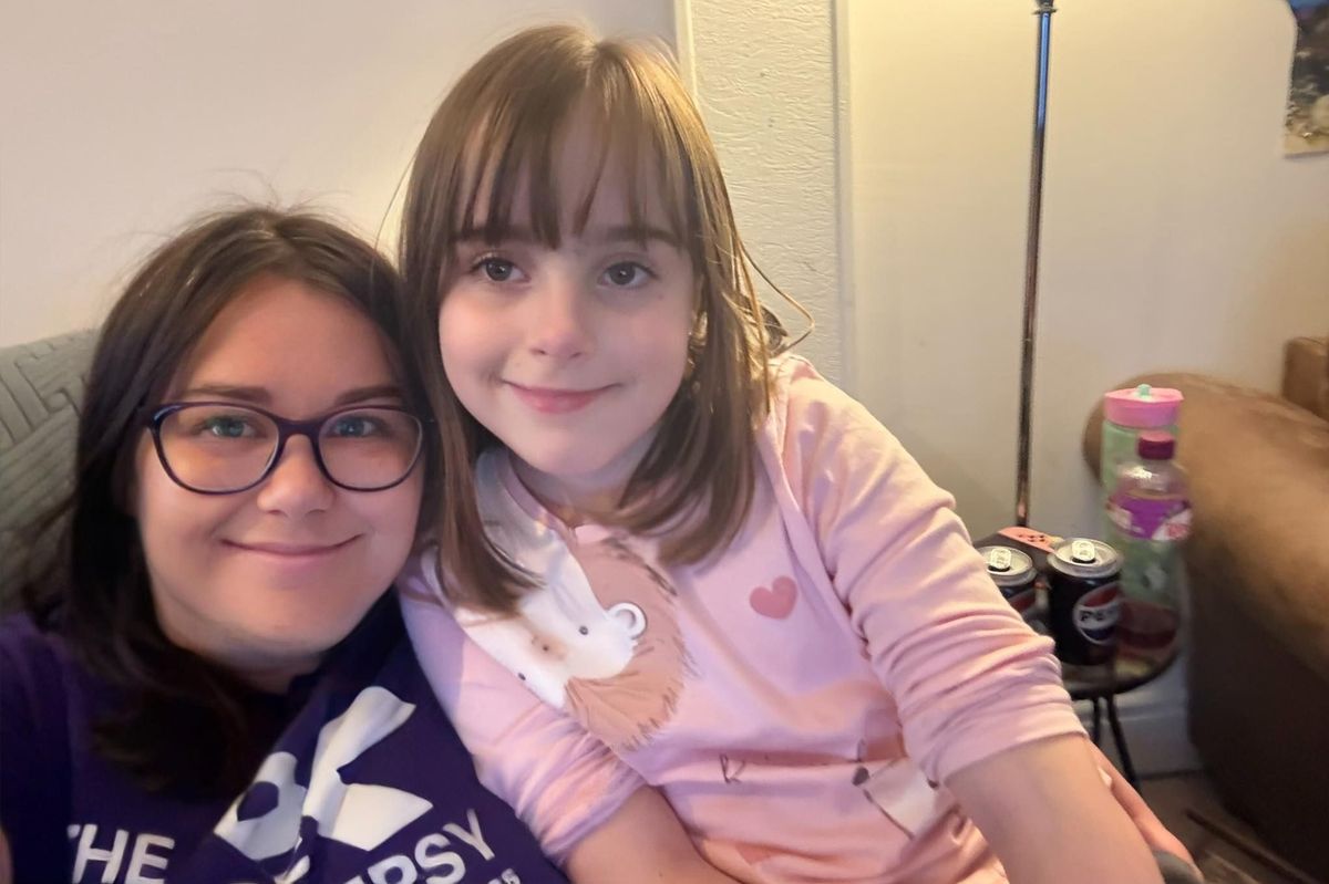 A young woman with dark hair and glasses, sat closely with a young 10 year old girl who has dark hair and a fringe - both are smiling