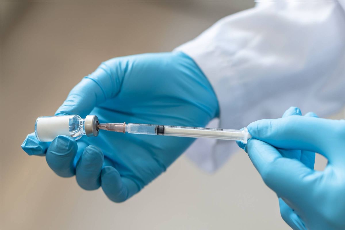 A person in a white lab coat and blue gloves is holding a syringe with a transparent needle, suggesting a medical or laboratory procedure.