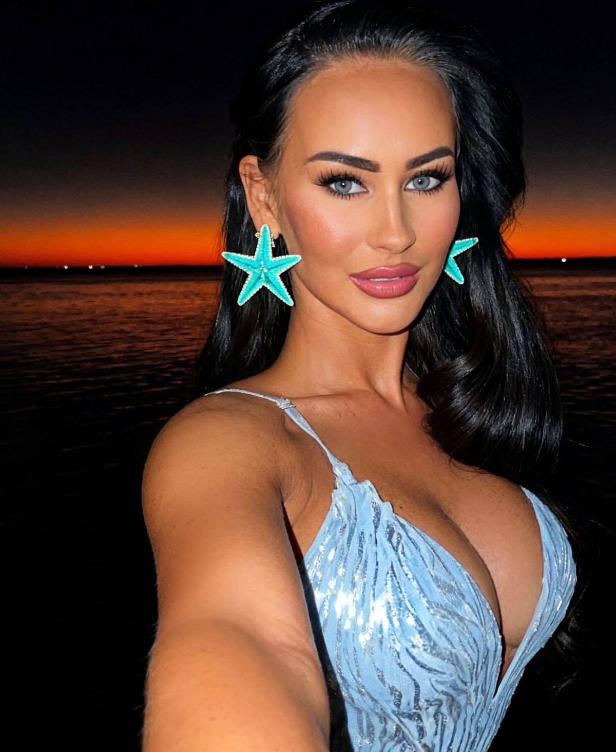 An image of a woman with long, dark hair and a serene expression. She is wearing turquoise starfish-shaped earrings and is standing in front of a serene twilight sky with hints of orange and pink. The background features a calm body of water, possibly a lake or sea, with a silhouette of a distant horizon.