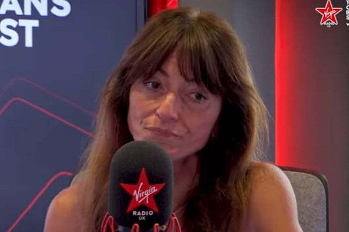 An individual with long hair is seated and speaking into a microphone adorned with a red star. The background features a blue and red graphic with the word "ST" prominently displayed.