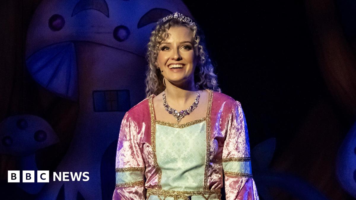 Alicia Belgarde wears a velvet pink and green dress. She has blonde curly hair and is wearing a tiara. She is stood on stage in front of a background which features mushrooms.