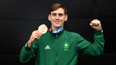 Sportsfile / Getty Images A man with short brown hair is smiling at the camera. He is holding one arm up and making a fist, in the other he is holding an Olympic medal. He is dressed in a green zip jacked with 'Adidas' and 'Tokyo 2020' branding. 
