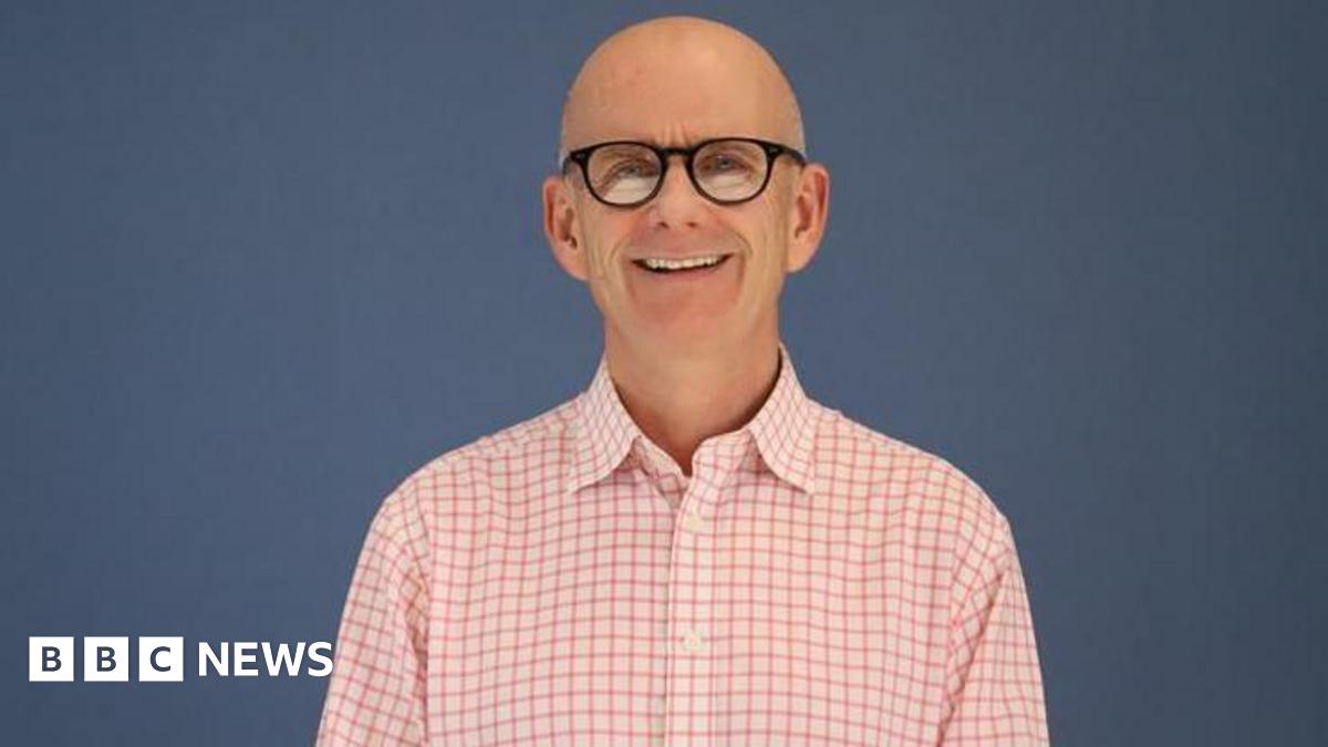 A man smiling at the camera. Here is wearing a pink chequered top and glasses. The background is blue.