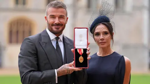 Getty Images Former England footballer David Beckham (L) poses next to his wife singer and fashion designer Victoria Beckham (R) with his medal after being appointed as a Knight Bachelor (Knighthood) for services to sport and charity at an investiture ceremony at Windsor Castle on November 4, 2025.