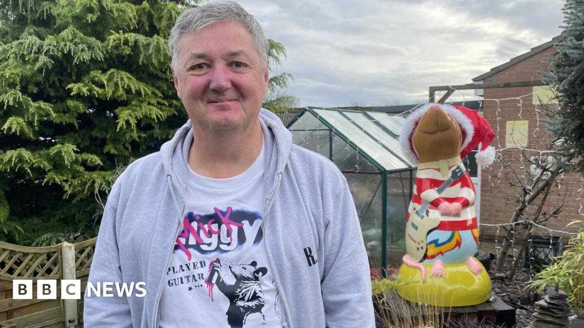 Jon Seth - a man wearing a grey hoodie and rat t-shirt honouring Mick Ronson - standing in front of a giant red and white striped rat sculpture holding a guitar. The sculpture has a red Santa hat on its head.