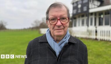 A man stood in a field with a building behind him which is black and white and surrounded by a fence and blurred. He has short grey hair, large red-rimmed glasses and a black blazer and light blue scarf on.