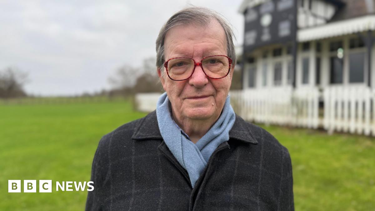 A man stood in a field with a building behind him which is black and white and surrounded by a fence and blurred. He has short grey hair, large red-rimmed glasses and a black blazer and light blue scarf on.