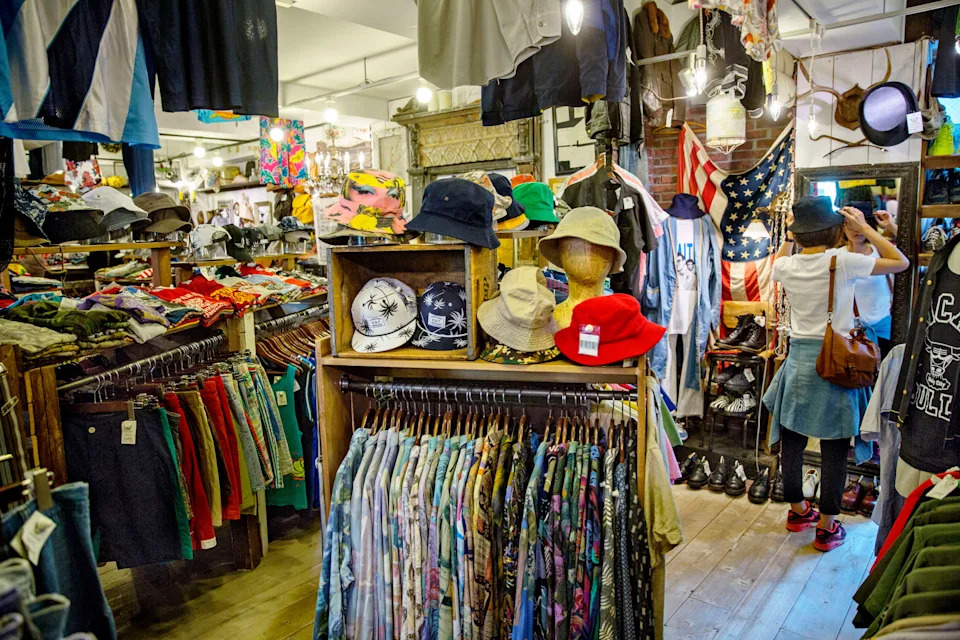 A thrift store interior filled with racks of diverse clothing and hats, with a person shopping, suggesting budget-friendly fashion choices