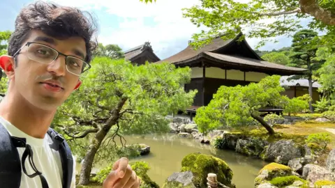 Ray Amjad Ray, who is wearing a white T-shirt and black rucksack, in front of a traditional Japanese garden with a pond.