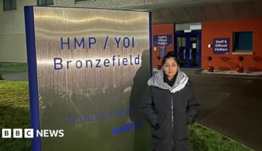 A woman in a long coat and black and white check scarf, standing outside a prison with her hands in her pockets