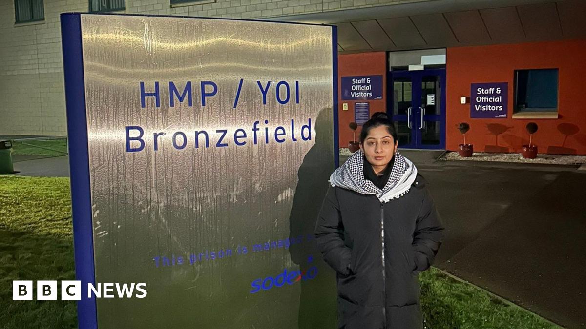 A woman in a long coat and black and white check scarf, standing outside a prison with her hands in her pockets